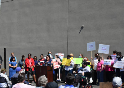 Photo of a group of people standing behind a woman speaking at a podium