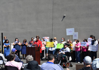 A group of people stand behind a woman speaking at a podium